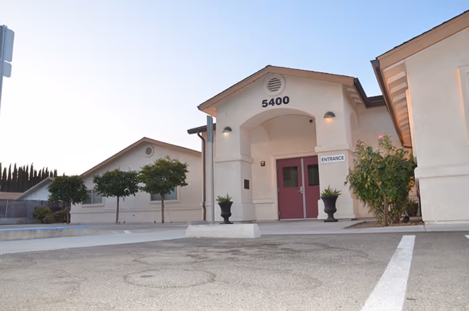 Front entrance of a light-colored assisted living building with the number 5400 above double doors and potted plants at the doorway.