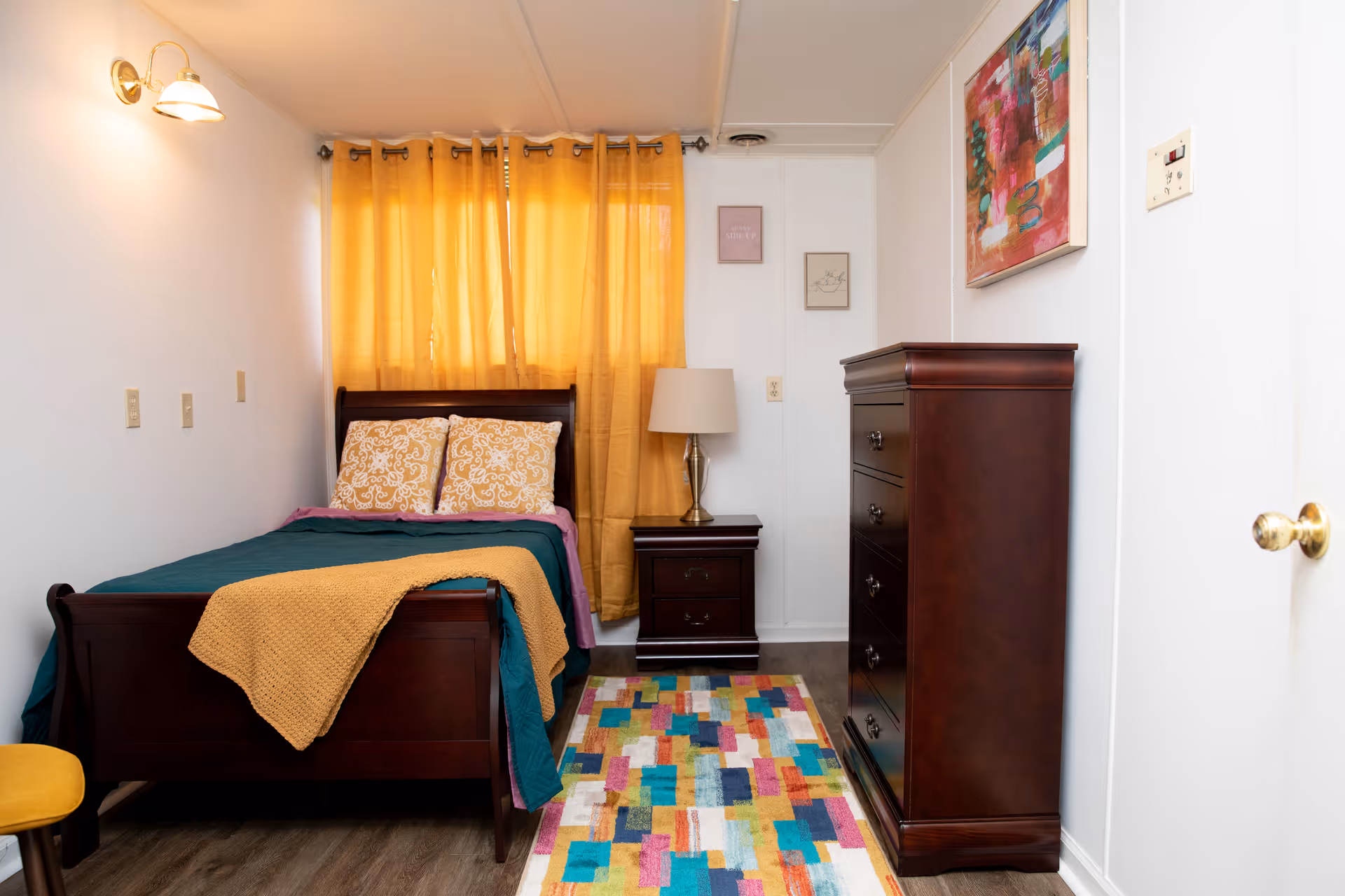 A cozy bedroom with a dark wooden bed frame, teal and mustard bedding, and two patterned pillows. There is a matching dark wooden nightstand with a lamp, a tall dresser, a colorful rectangular rug on the floor, mustard yellow curtains covering the window, and two small framed pictures on the wall.