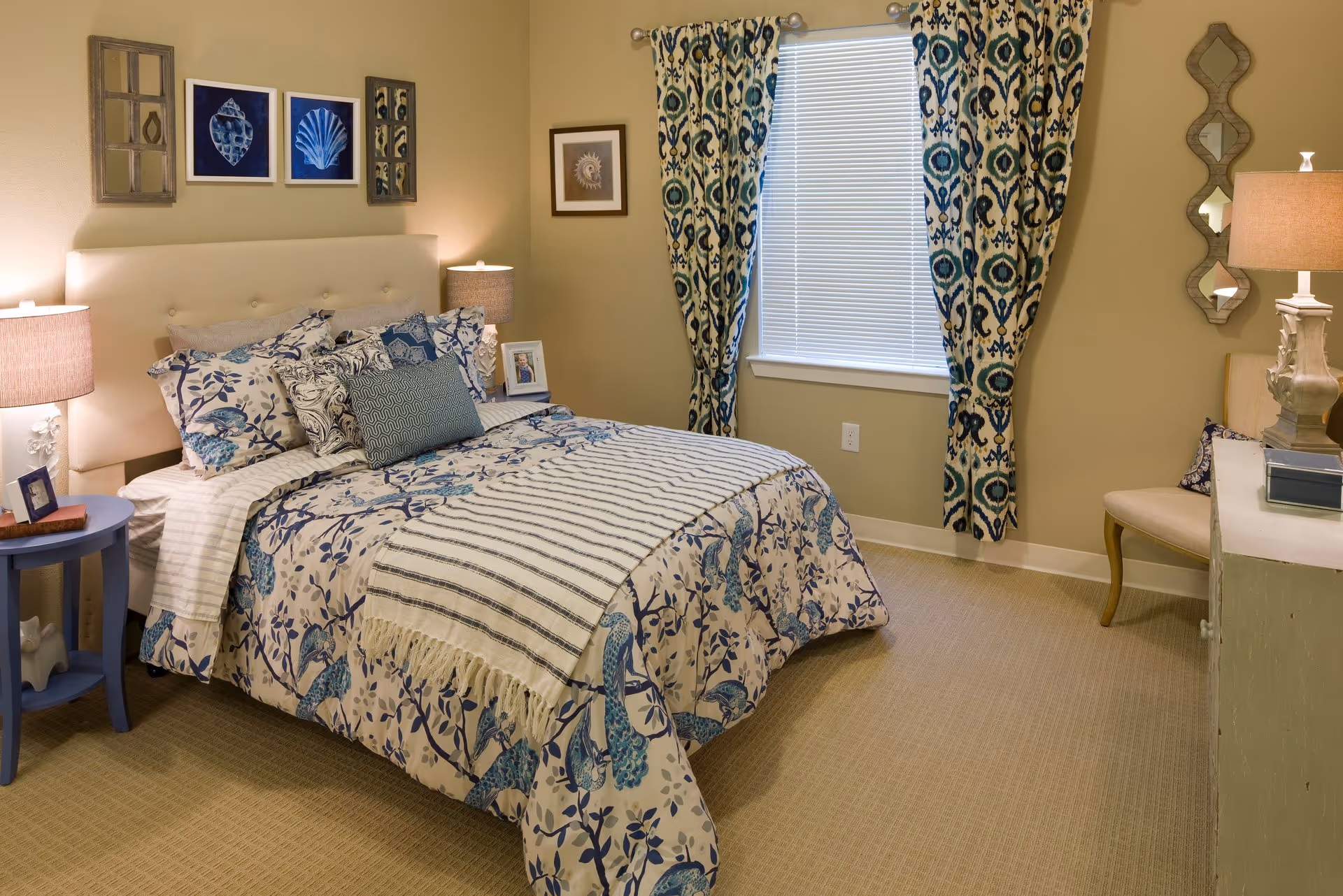 Well-decorated bedroom with a bed dressed in blue patterned bedding, bedside tables and lamps, patterned curtains over a window, and wall art.