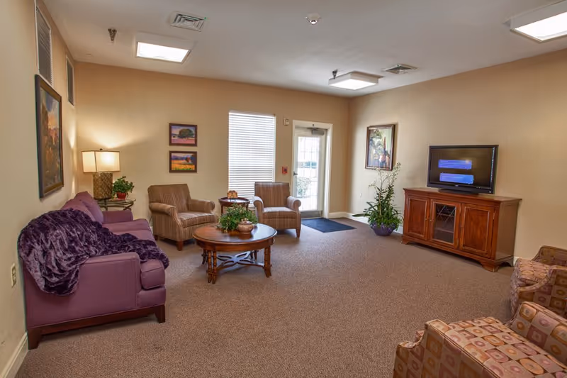 A cozy senior living facility common area with a purple sofa, two striped armchairs, a wooden coffee table with plants, a wooden TV stand with a flat-screen TV, and additional patterned armchairs. The room has beige walls, carpeted floor, framed artwork, a table lamp, and a glass door letting in natural light.