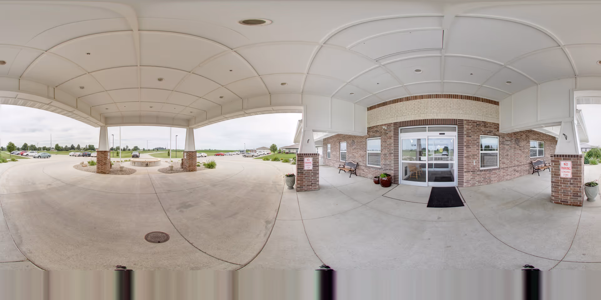Covered drive-up entrance with a canopy in front of a brick senior living facility, showing glass double doors, benches, planters, and parked cars in the background.