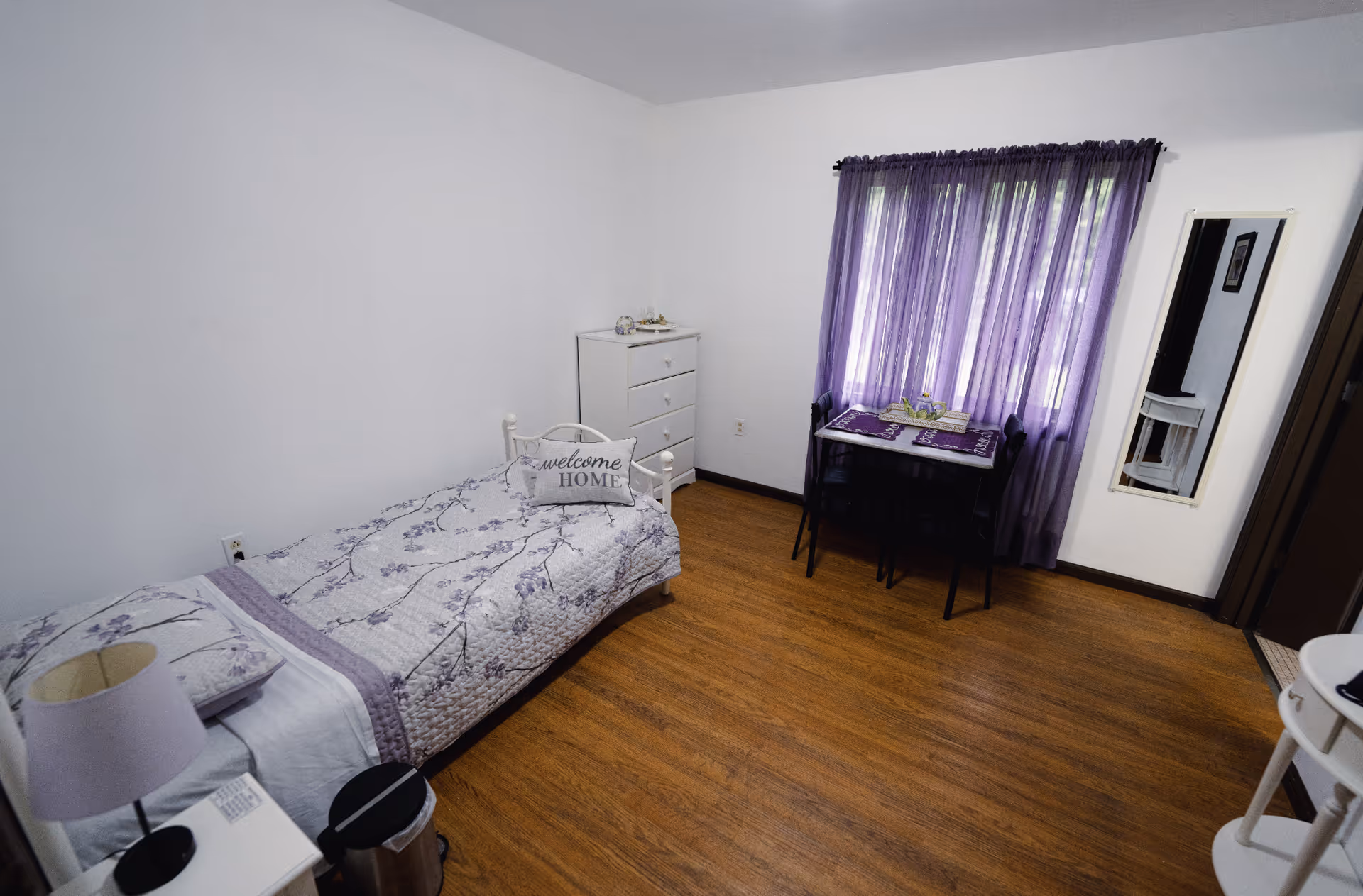 Small bedroom with a single bed in purple floral bedding, a white dresser, a small table by a window with purple curtains, and wood flooring.