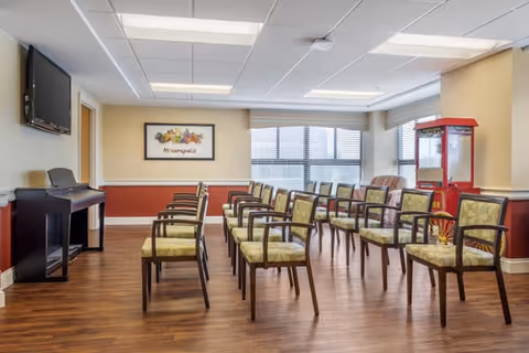 A well-lit room with rows of wooden chairs featuring green patterned cushions arranged facing a wall-mounted flat screen TV. The room has wood flooring, beige and red walls, large windows with blinds, a framed artwork on the wall, and a red popcorn machine in the corner.