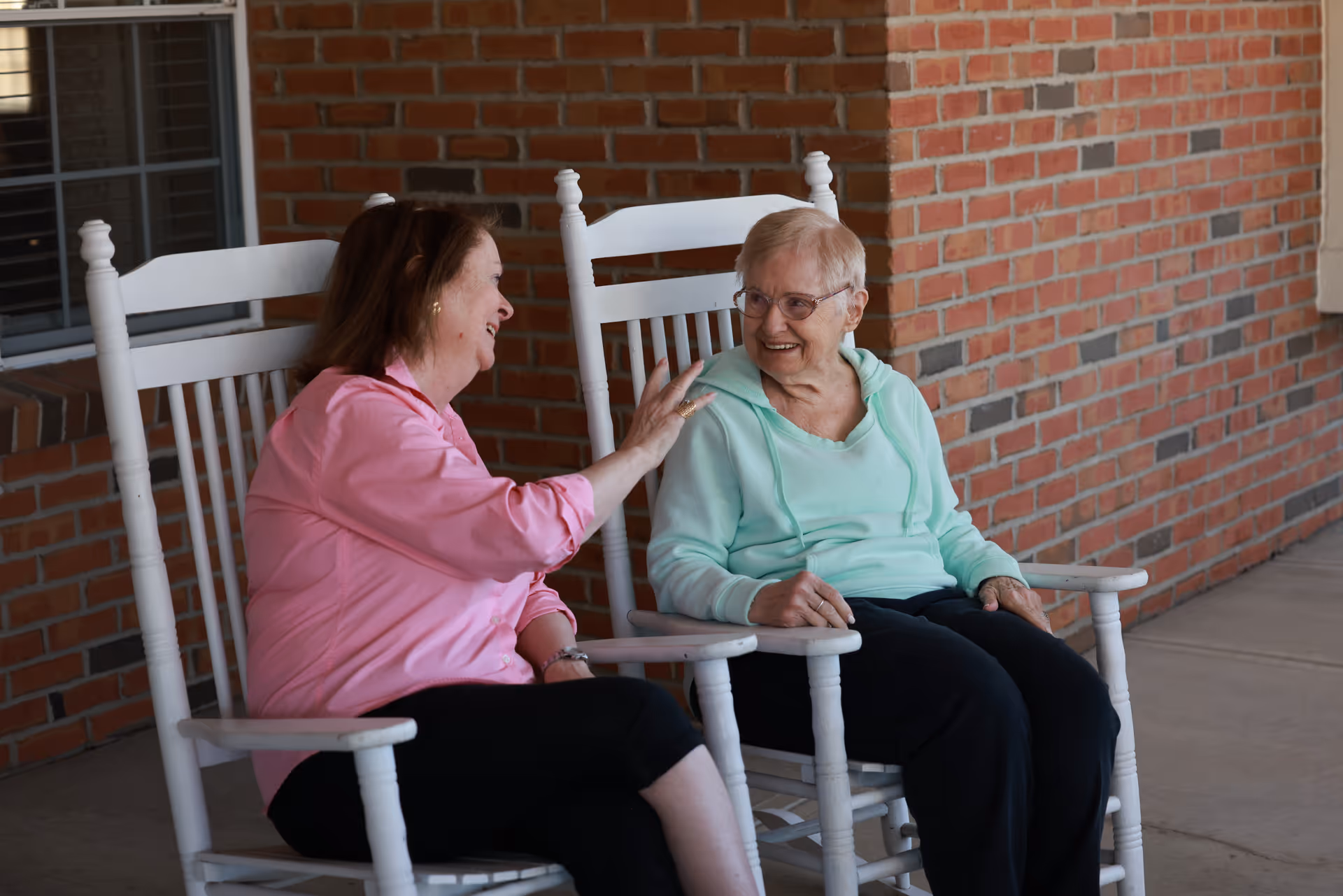 Two elderly women sitting and chatting happily on white rocking chairs on a covered porch with a brick wall background.