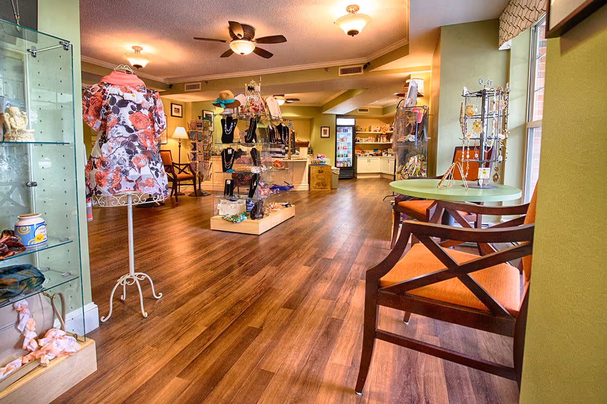 Interior view of a retail or gift shop area in a senior living facility with wooden flooring, display shelves and racks holding jewelry, hats, and clothing. There are chairs with orange cushions and a small round table near a window on the right side. The walls are painted green and the ceiling has multiple light fixtures and ceiling fans.