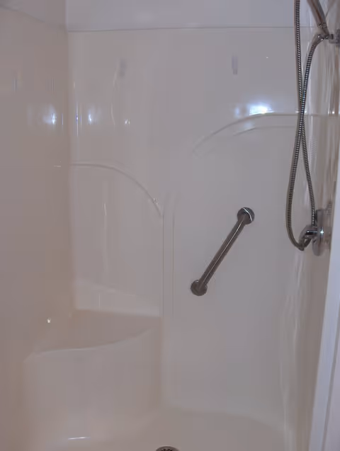A white shower stall with a built-in corner seat, a horizontal grab bar on the wall, and a handheld showerhead attached to a flexible hose.