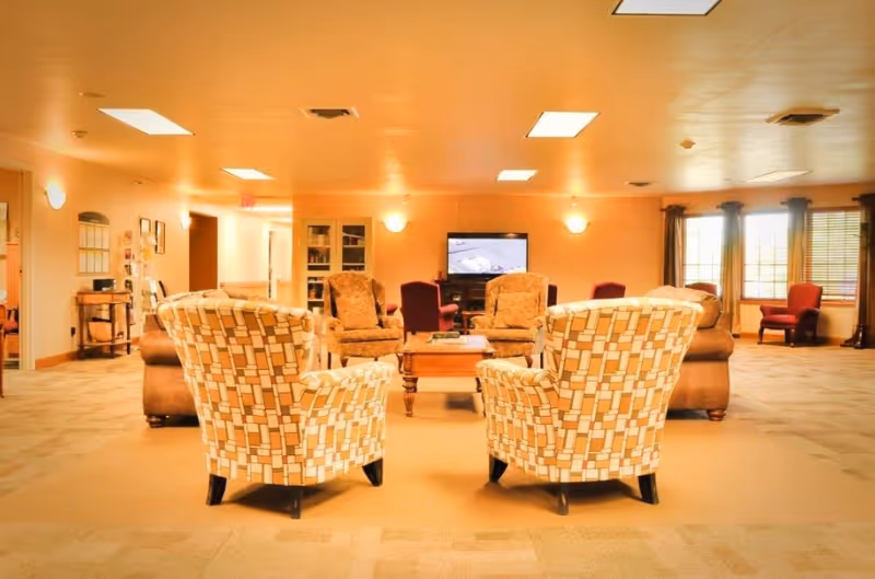 A spacious living room area with patterned armchairs in the foreground, two upholstered armchairs and a wooden coffee table in the center, a TV mounted on the wall, and large windows with curtains letting in natural light.