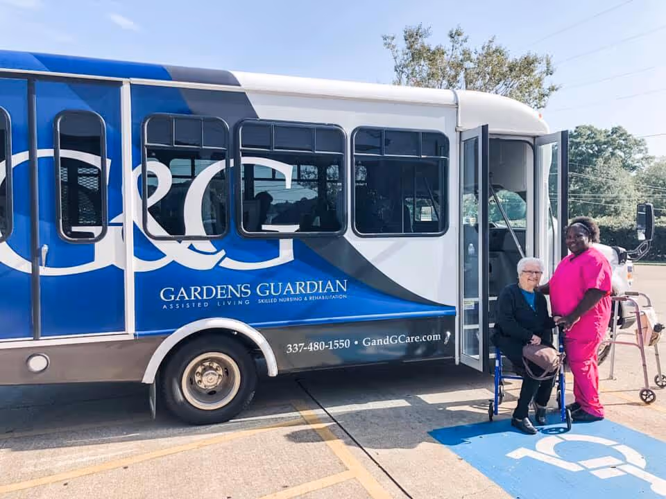 A senior woman sitting in a wheelchair next to a caregiver dressed in pink scrubs outside a large blue and white shuttle bus with the text 'Gardens Guardian Assisted Living Skilled Nursing & Rehabilitation' on its side. They are in a parking lot with a handicap parking space visible.