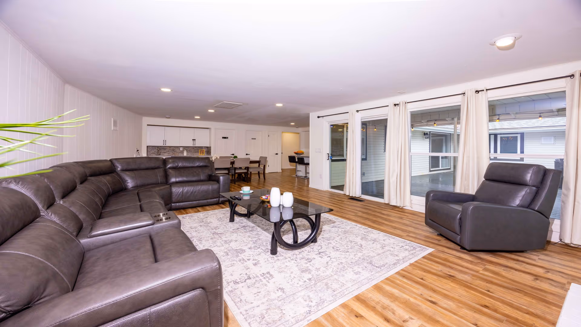 Spacious living room with a large dark brown leather sectional sofa and a matching armchair. A glass coffee table sits on a patterned area rug over wooden flooring. The room has white walls, recessed lighting, and large windows with white curtains that look out onto an outdoor covered patio area. In the background, there is a dining area with a table and chairs and a small kitchenette with cabinets.