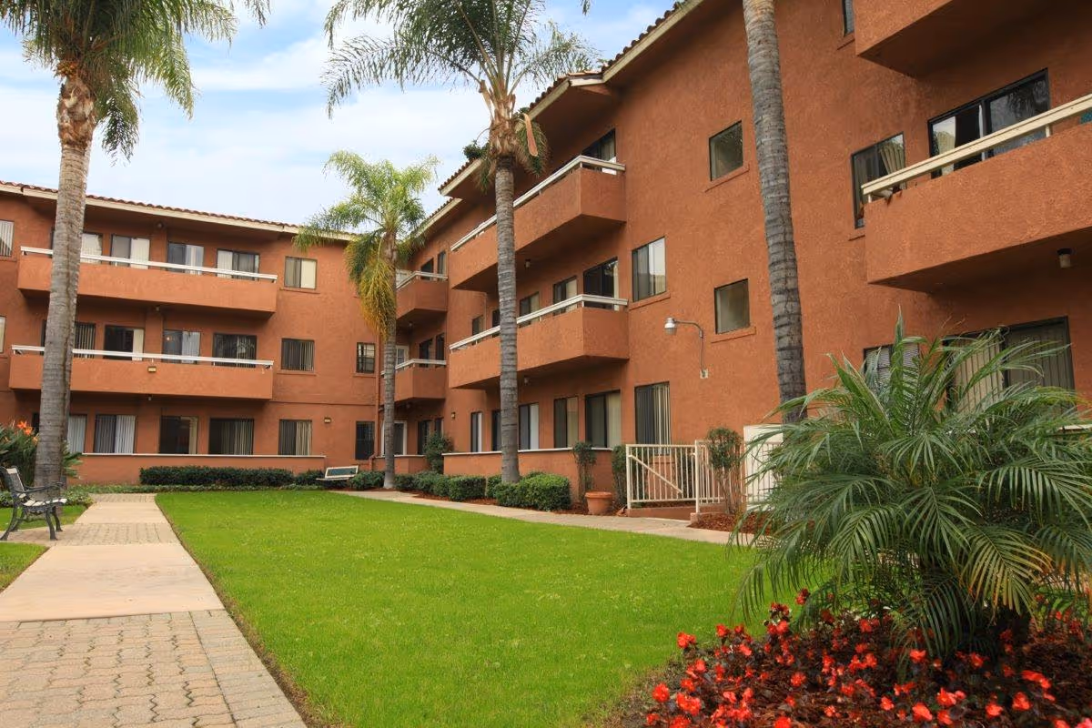 Outdoor courtyard area of a senior living facility with green grass, palm trees, flower beds with red flowers, benches, and a three-story building with balconies and windows in the background.