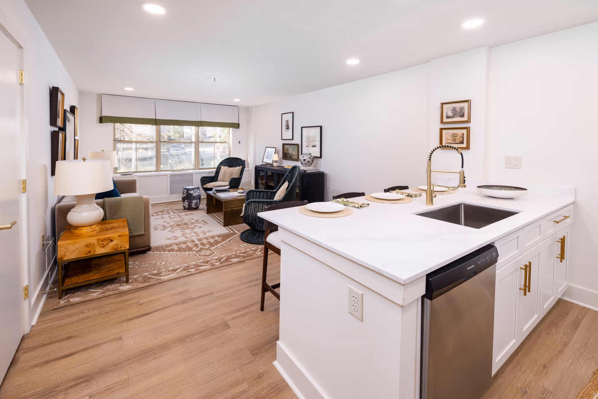 Bright and modern open-concept living space featuring a kitchen island with a sink and four place settings, adjacent to a cozy living room area with a sofa, two wicker chairs, a wooden coffee table, and large windows letting in natural light.