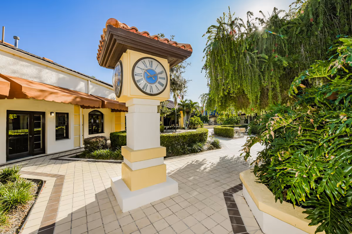 Outdoor courtyard area at The Barclay at Sarasota featuring a decorative clock on a pedestal with Roman numerals, surrounded by tiled walkways, lush green plants, and a building with a terracotta awning under a clear blue sky.