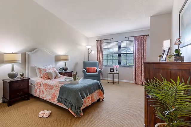 A cozy bedroom with a single bed covered in floral bedding and a green throw blanket. There are two bedside tables with lamps on either side of the bed. A comfortable armchair with a red cushion is placed near a window with pink patterned curtains. A small side table with flowers and a photo frame is next to the chair. A wooden dresser with plants and framed pictures is on the right side of the room. The floor is carpeted and a pair of pink slippers are placed near the bed.