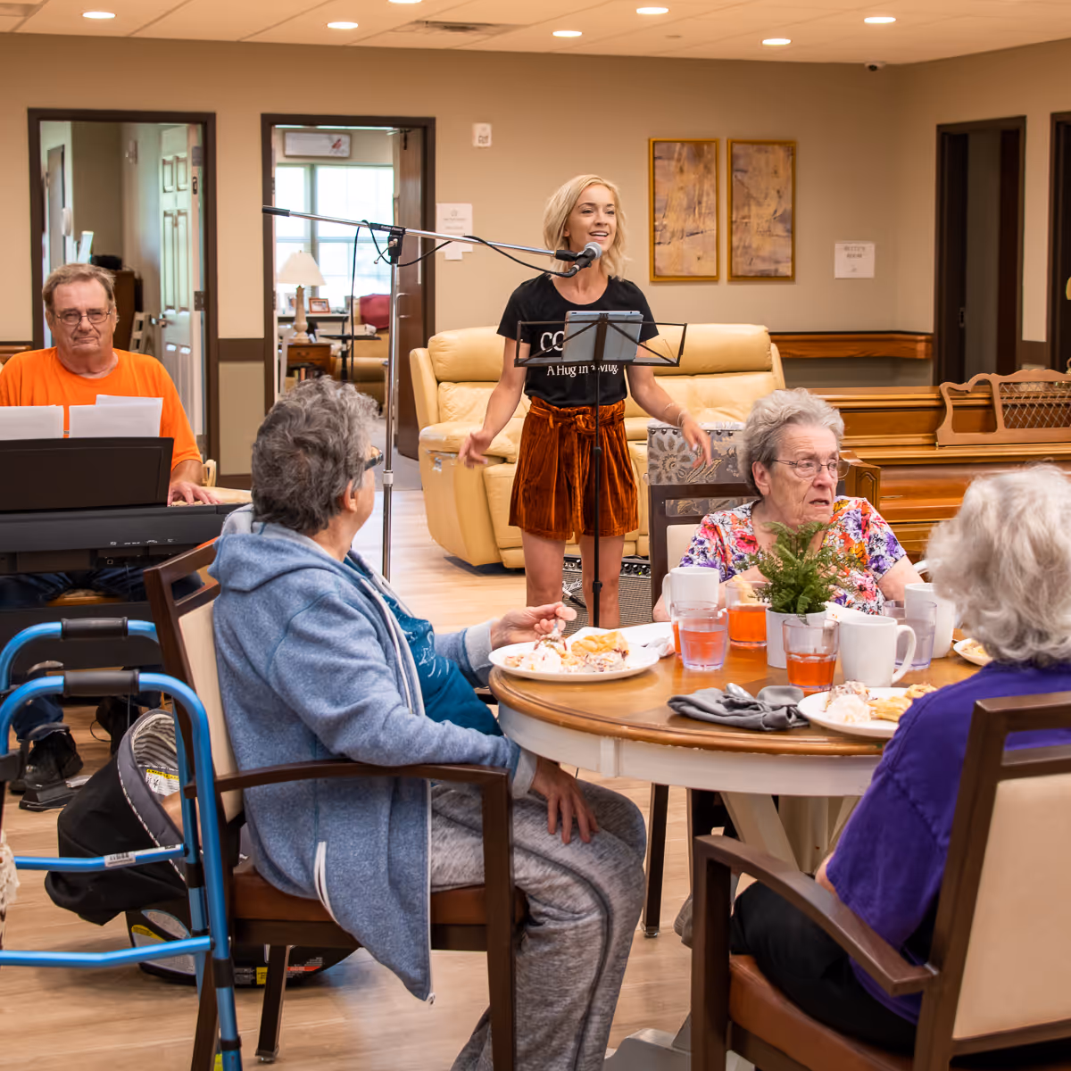 A woman sings into a microphone while seniors eat at a round table and a man plays keyboard in a senior living dining/activity room.