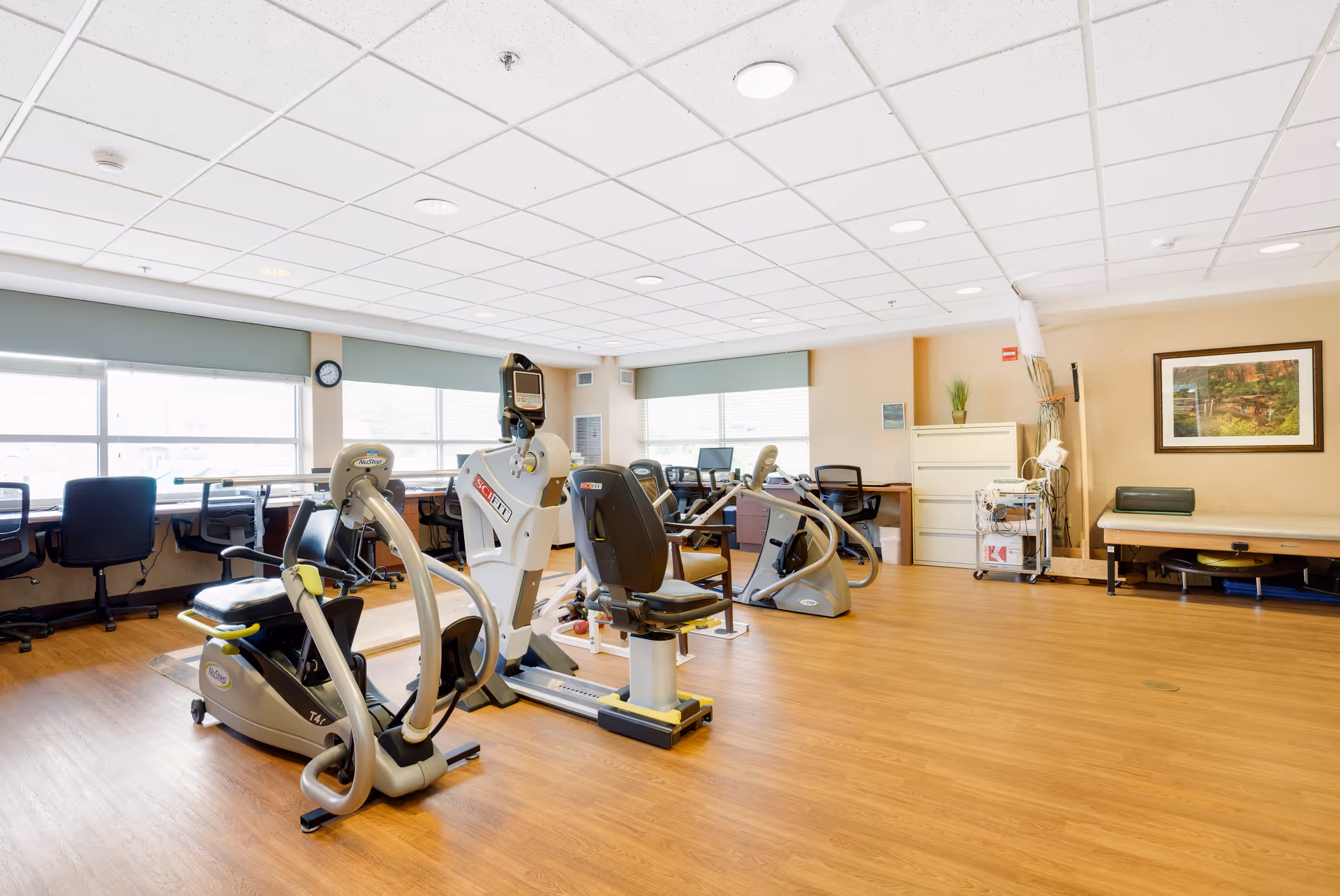A bright and spacious exercise room in an assisted living facility with several pieces of fitness equipment including recumbent bikes and elliptical machines. The room has large windows with green blinds, a wooden floor, and a ceiling with recessed lighting. There are chairs and desks along the windows, a filing cabinet, a treatment table, and a framed picture on the wall.