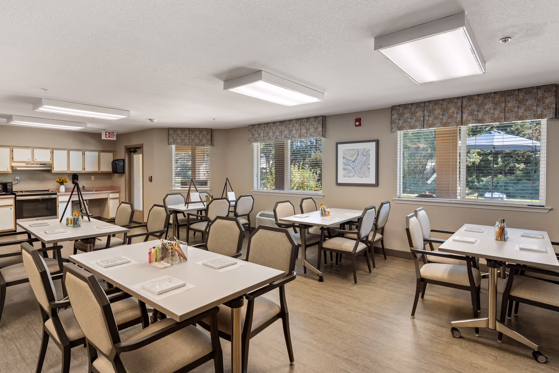 A bright dining room in a senior living community with several tables and chairs arranged neatly. Each table is set with napkins and condiment holders. Large windows with patterned valances allow natural light to fill the room, and a small kitchen area with cabinets and appliances is visible in the background.