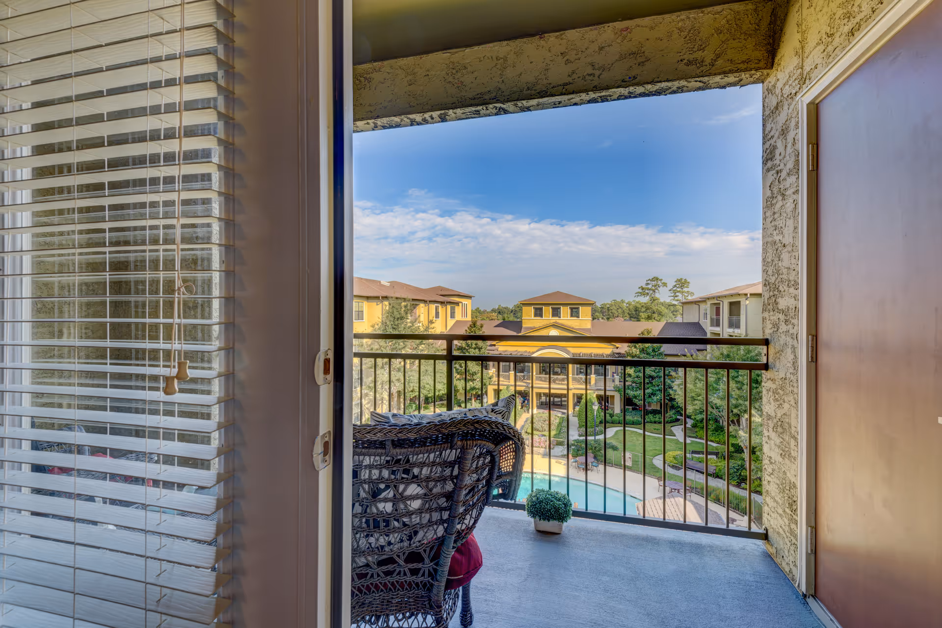 View from a balcony with a wicker chair and a small potted plant, overlooking a swimming pool and landscaped courtyard surrounded by multi-story buildings under a partly cloudy blue sky.