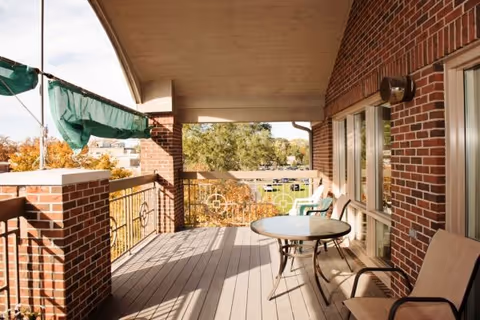 Covered outdoor balcony with a round table and chairs, brick walls, and a view of trees and nearby buildings.