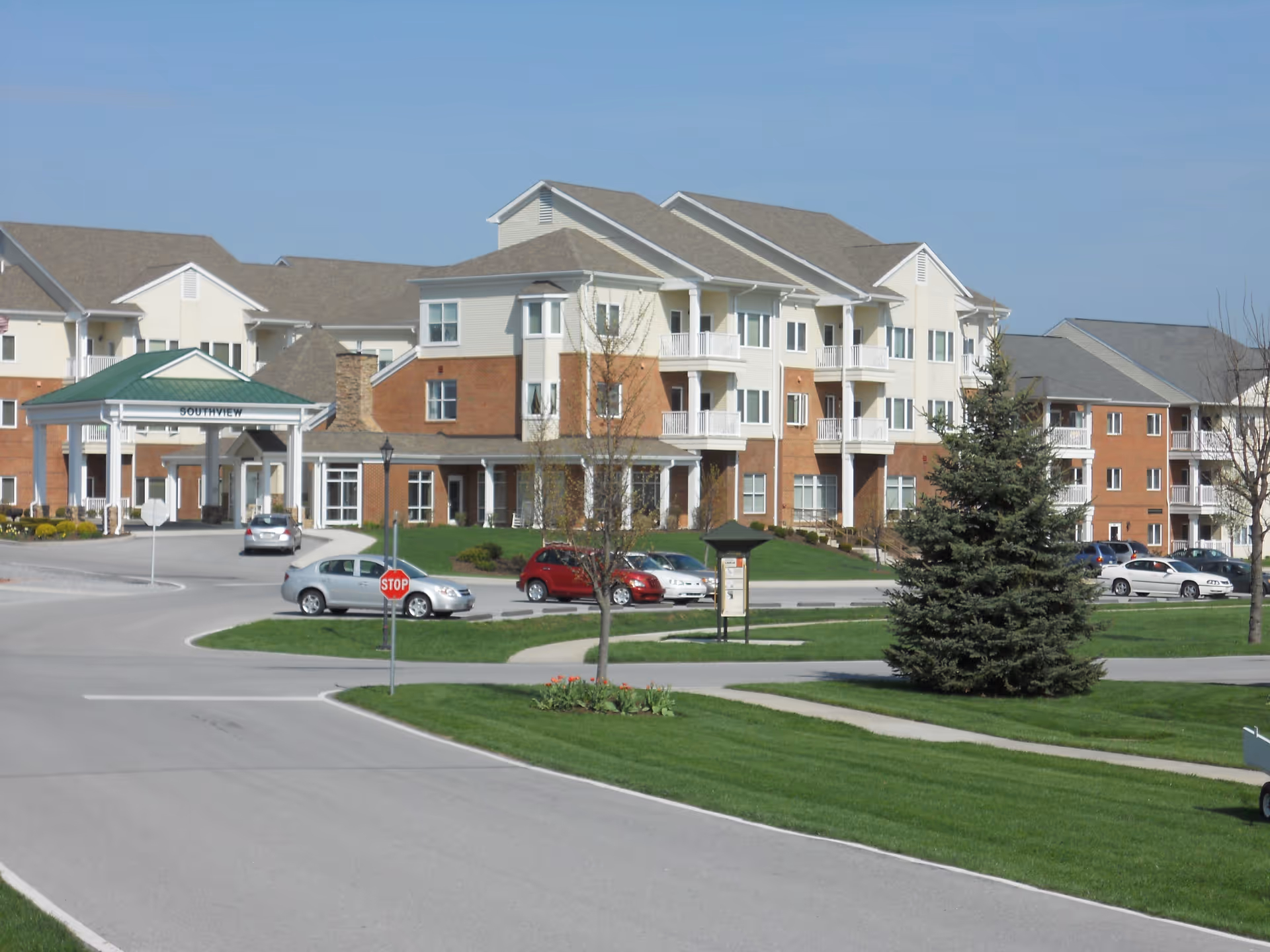 Three-story senior living building with a covered entrance, parked cars and landscaped lawns under a clear sky.