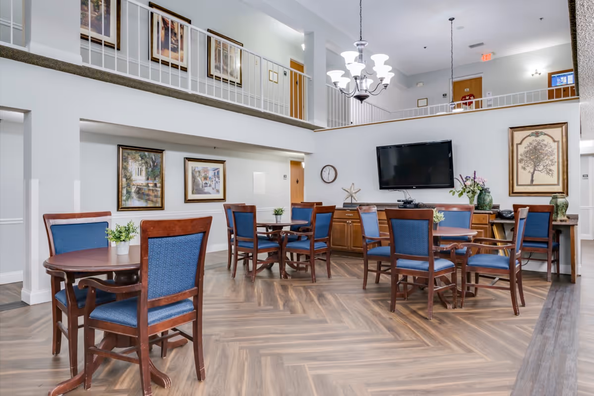 A spacious common area with multiple round wooden tables and blue upholstered chairs arranged around them. The room features a high ceiling with a chandelier, a large flat-screen TV mounted on the wall, framed artwork, and decorative plants. There is a balcony railing on the upper level overlooking the area, and the floor has a wood pattern design.