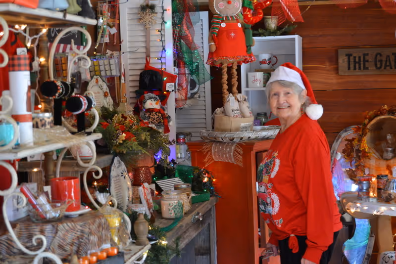 An elderly woman wearing a red Christmas sweater and a Santa hat stands smiling in a room decorated with various holiday ornaments, lights, and festive decorations including a gingerbread doll, wreaths, and candles.