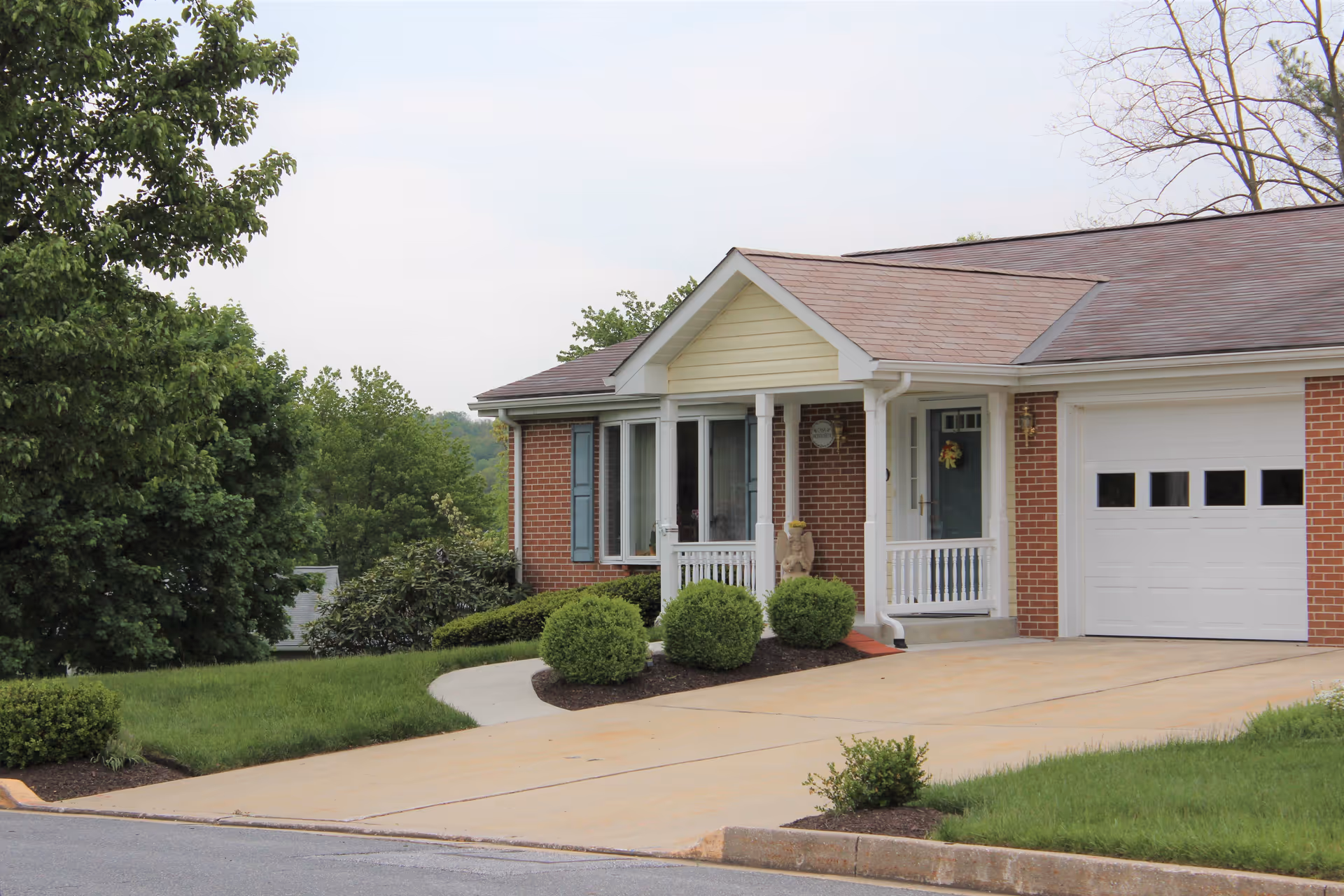 Exterior view of a single-story brick house with a small covered porch, white railings, a blue front door with a wreath, and an attached white garage door. The house is surrounded by green bushes, a well-maintained lawn, and trees in the background under a cloudy sky.