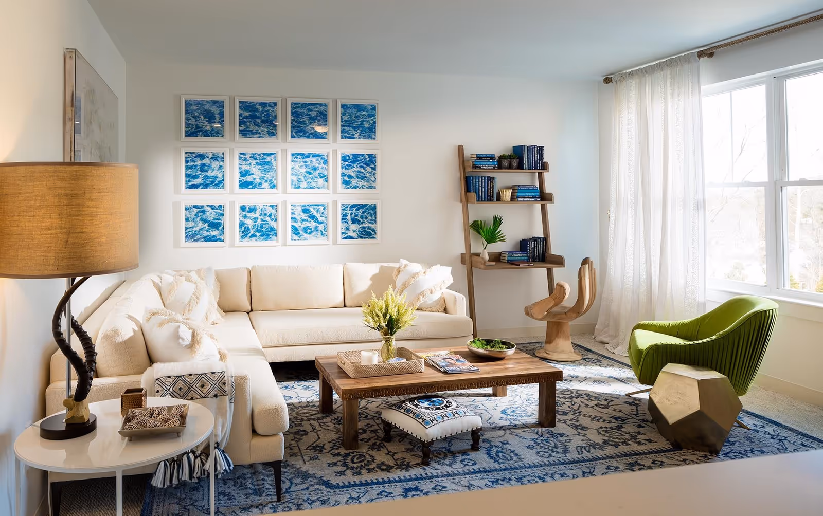 Bright and cozy living room with a large beige sectional sofa adorned with white pillows and a throw blanket. A wooden coffee table with a vase of yellow flowers, magazines, and a tray sits on a blue patterned rug. To the right, a unique wooden hand-shaped chair and a green upholstered chair are positioned near a large window with sheer white curtains. A wooden ladder-style bookshelf with books and plants is against the wall, and a large lamp with a beige shade is on a small round side table next to the sofa. Above the sofa, a grid of framed pictures depicting water scenes is displayed on the wall.