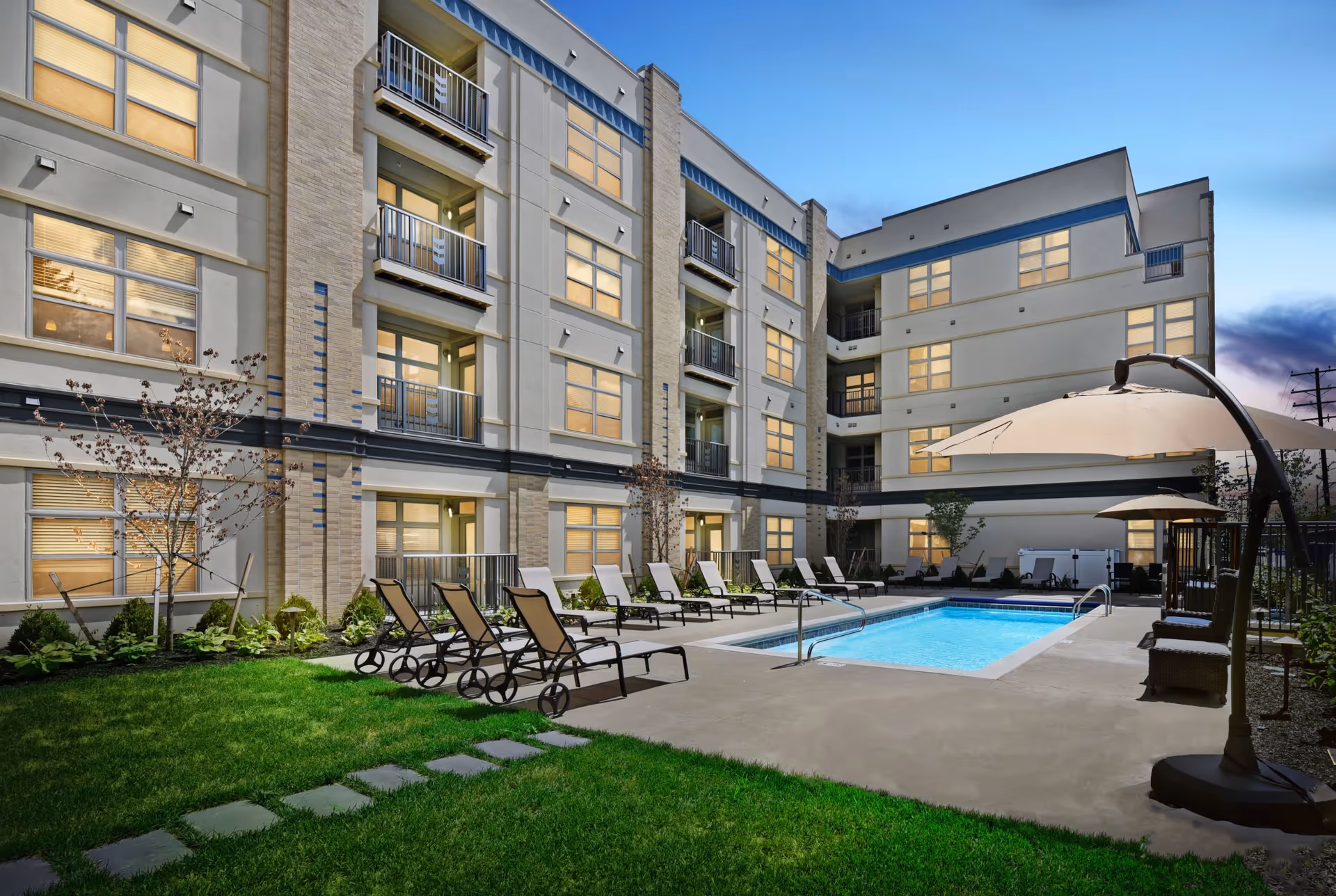 Outdoor courtyard area of The Deco facility at dusk featuring a rectangular swimming pool surrounded by lounge chairs and large umbrellas, with a well-maintained grassy area and a multi-story building with balconies and lit windows in the background.