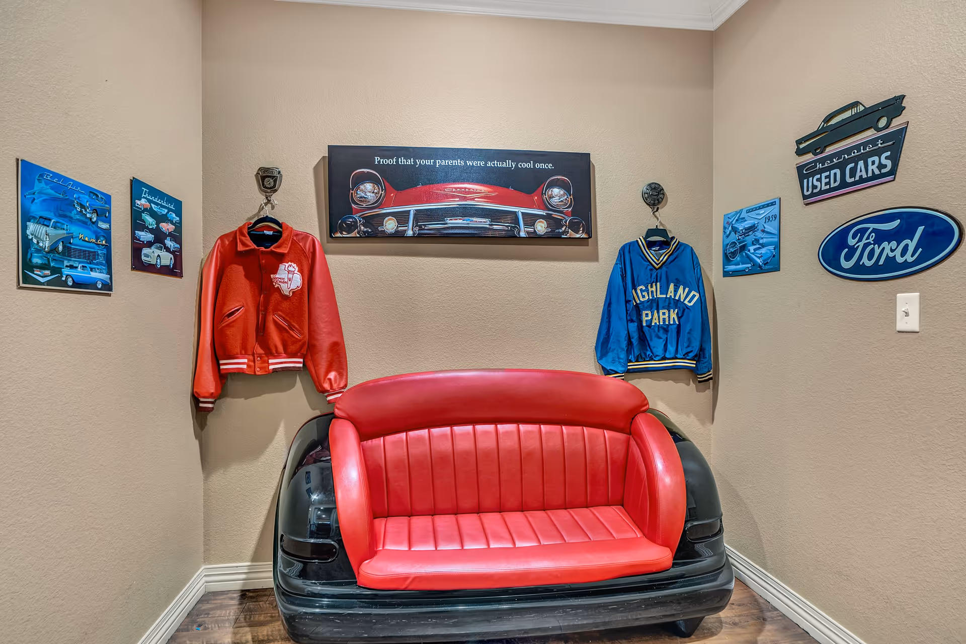 A small corner room with beige walls featuring a red and black car-themed couch resembling the back of a vintage car. Above the couch is a framed picture of a classic red car with the text 'Proof that your parents were actually cool once.' On the left wall, two vintage car posters and a red jacket are hanging. On the right wall, a blue jacket, a Chevrolet Used Cars sign, a Ford logo sign, and a vintage car poster are displayed. The floor is wooden.