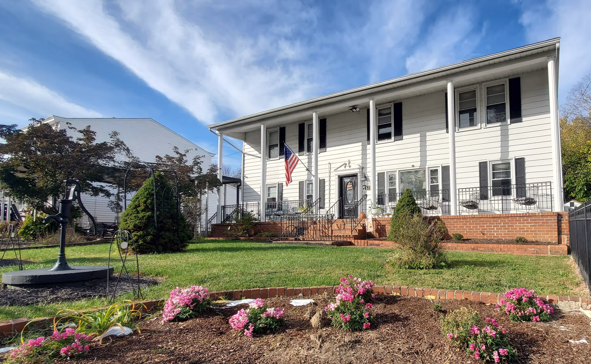 Front exterior view of a two-story white building with black shutters and a black front door, an American flag hanging near the entrance, a well-maintained lawn, flower beds with pink flowers, and a black water pump in the garden area under a partly cloudy blue sky.
