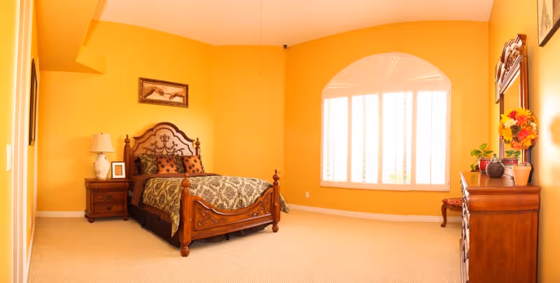A bedroom with bright yellow walls featuring a wooden bed with an ornate headboard and patterned bedding. There is a wooden nightstand with a lamp and framed pictures on the left side of the bed. A large arched window with white shutters allows natural light into the room. On the right side, there is a wooden dresser with a mirror, decorated with a vase of flowers and other small items.