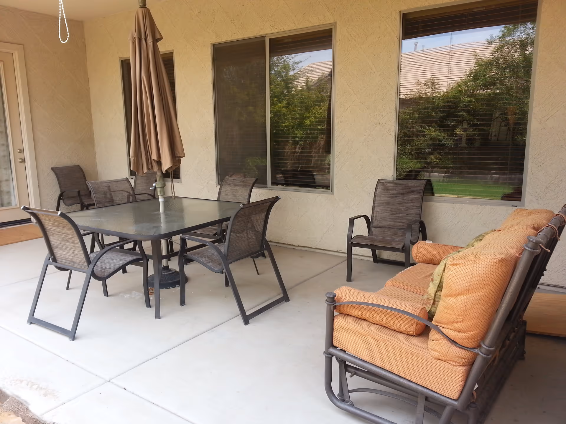 Outdoor patio area with a glass-top table surrounded by six metal chairs with mesh seats and backs. There is a large closed beige umbrella in the center of the table. To the right, there is a cushioned metal bench with orange cushions and pillows. The patio is adjacent to a building with two large windows reflecting greenery outside.