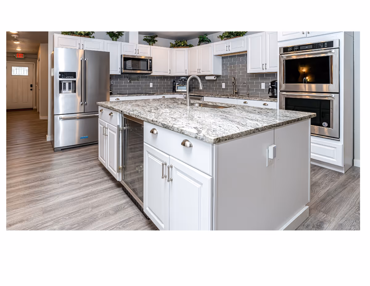 Modern kitchen with white cabinetry, a large granite island with a built-in sink and faucet, stainless steel refrigerator, microwave, double oven, and a gray subway tile backsplash. The floor is light wood, and there are plants on top of the cabinets.
