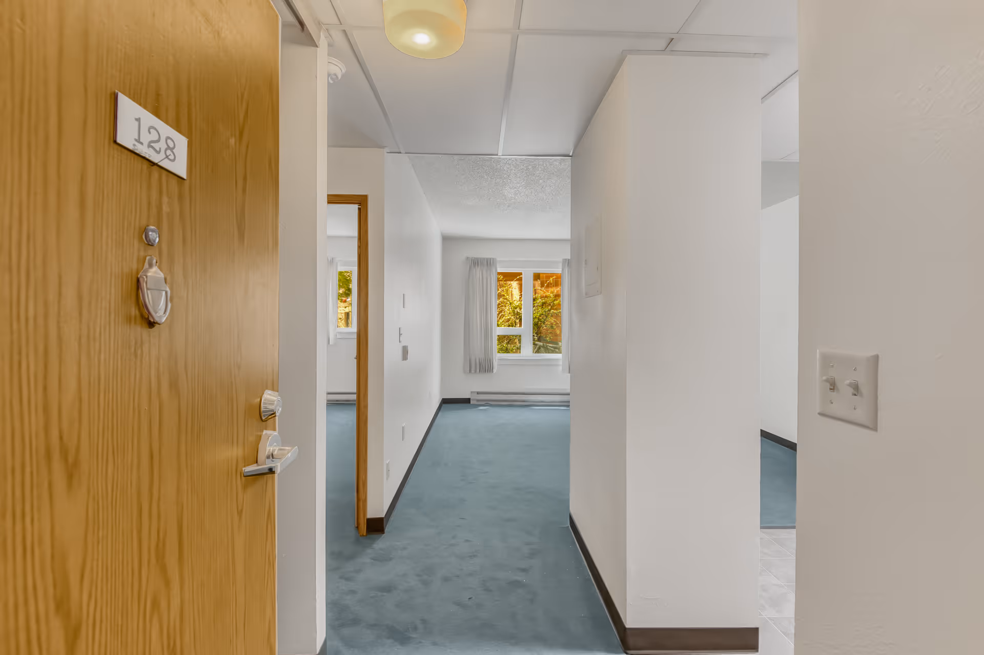 View of an empty apartment hallway with a wooden door labeled 128 on the left, leading into a carpeted room with white walls and a window with curtains letting in natural light.