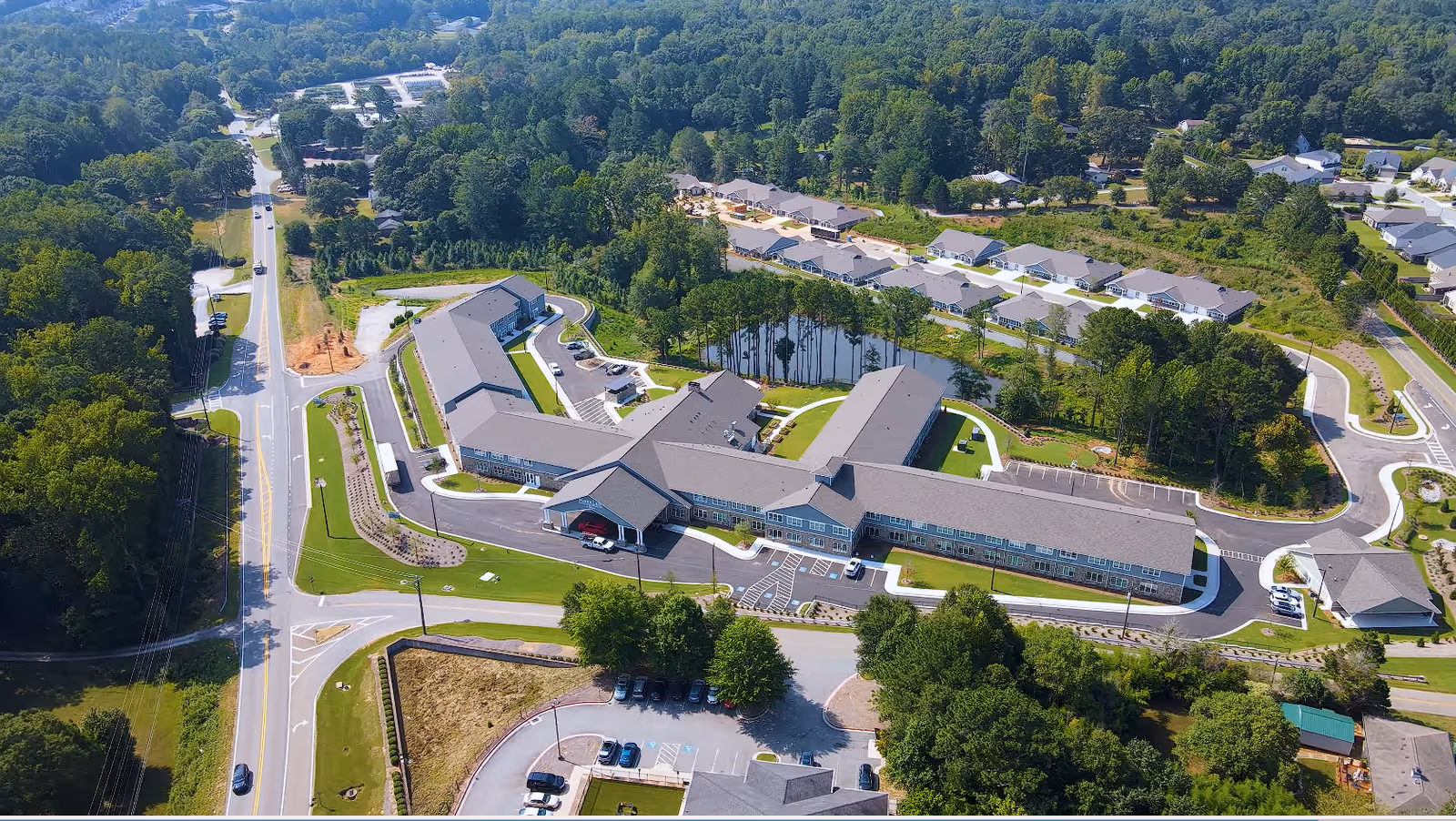 Aerial view of Manor Lake Assisted Living, Memory Care & Independent Living facility in Hickory Flat, showing a large building complex with multiple wings surrounded by greenery, parking lots, roads, and nearby residential houses.