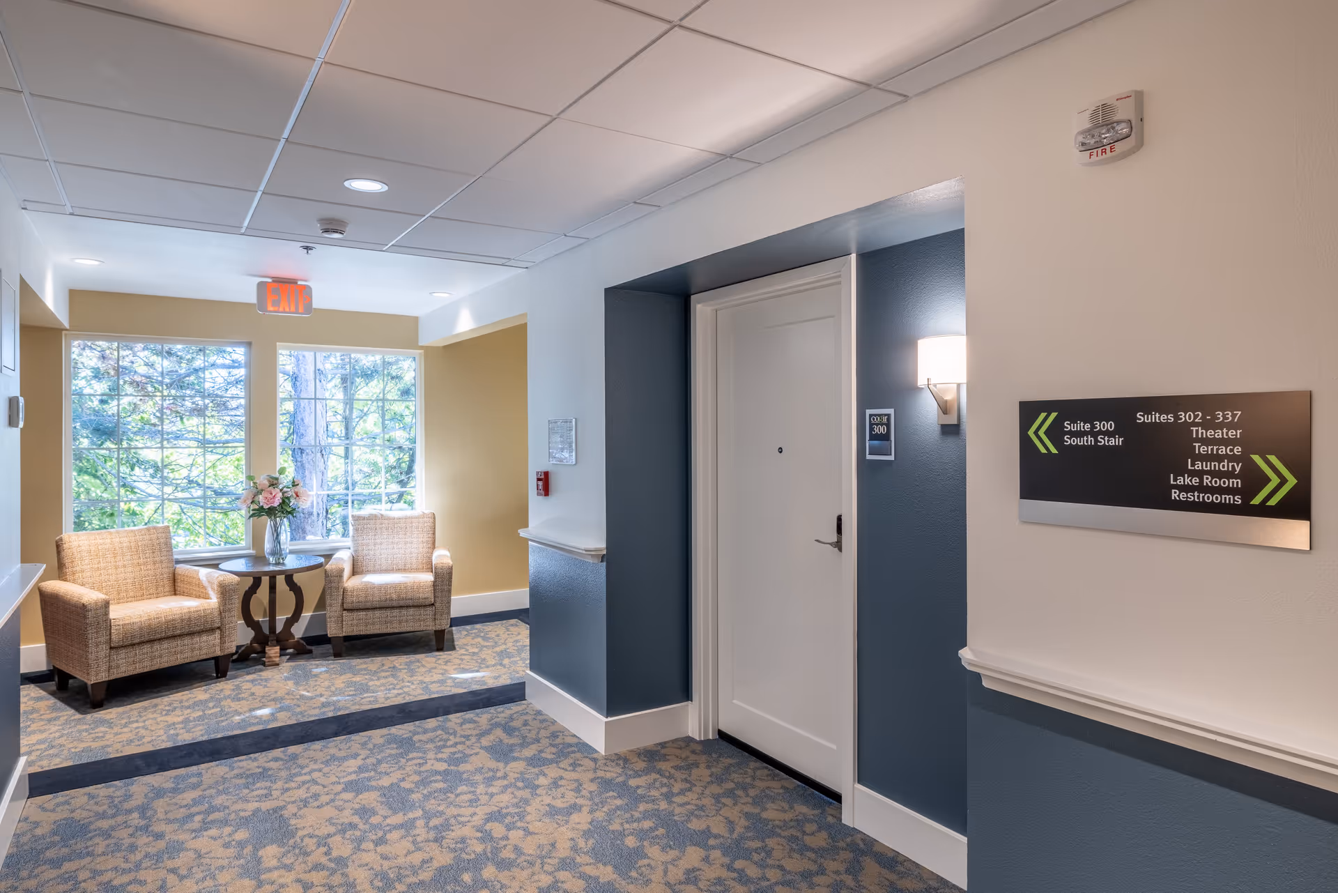 A well-lit hallway in a senior living facility with a seating area by a large window. The seating area has two cushioned armchairs and a small round table with a vase of flowers. There is a white door labeled Suite 300, a wall-mounted light, and a directional sign indicating locations such as suites, theater, terrace, laundry, lake room, and restrooms.