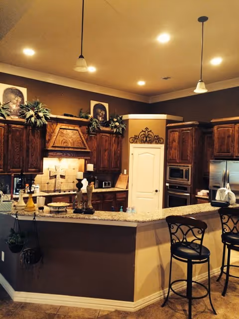 A warm-toned kitchen interior featuring wooden cabinets, a granite countertop island with decorative items, two high chairs with ornate backs, stainless steel appliances including a refrigerator and built-in microwave, and recessed ceiling lights. The kitchen has a cozy and inviting atmosphere with plants and artwork above the cabinets.
