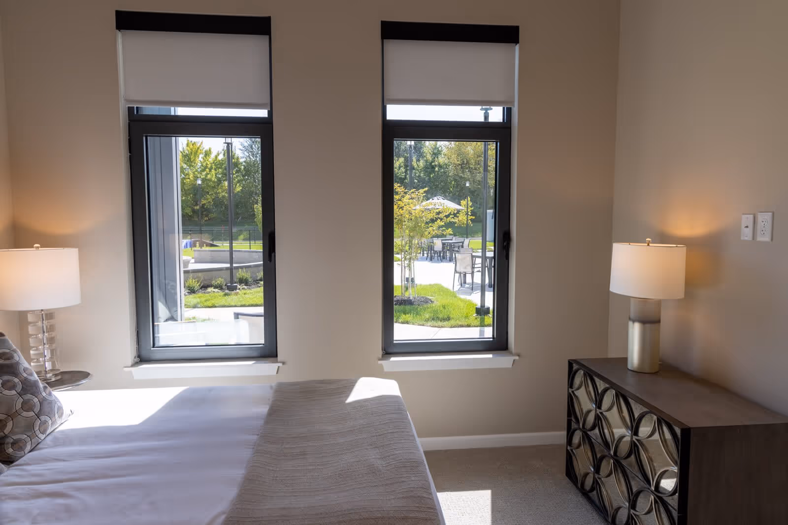 Sunlit bedroom interior with a bed, two bedside lamps, a dresser and two windows looking out to an outdoor patio.