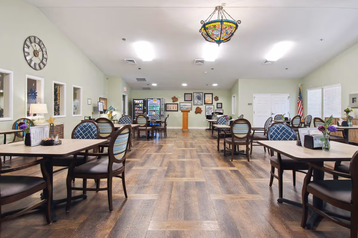 A spacious dining room with multiple tables and chairs arranged neatly. The room has wooden flooring, light green walls, and a large decorative clock on the left wall. There is a colorful stained glass chandelier hanging from the ceiling. At the far end, there are vending machines and framed pictures on the wall. An American flag is visible near the windows on the right side.