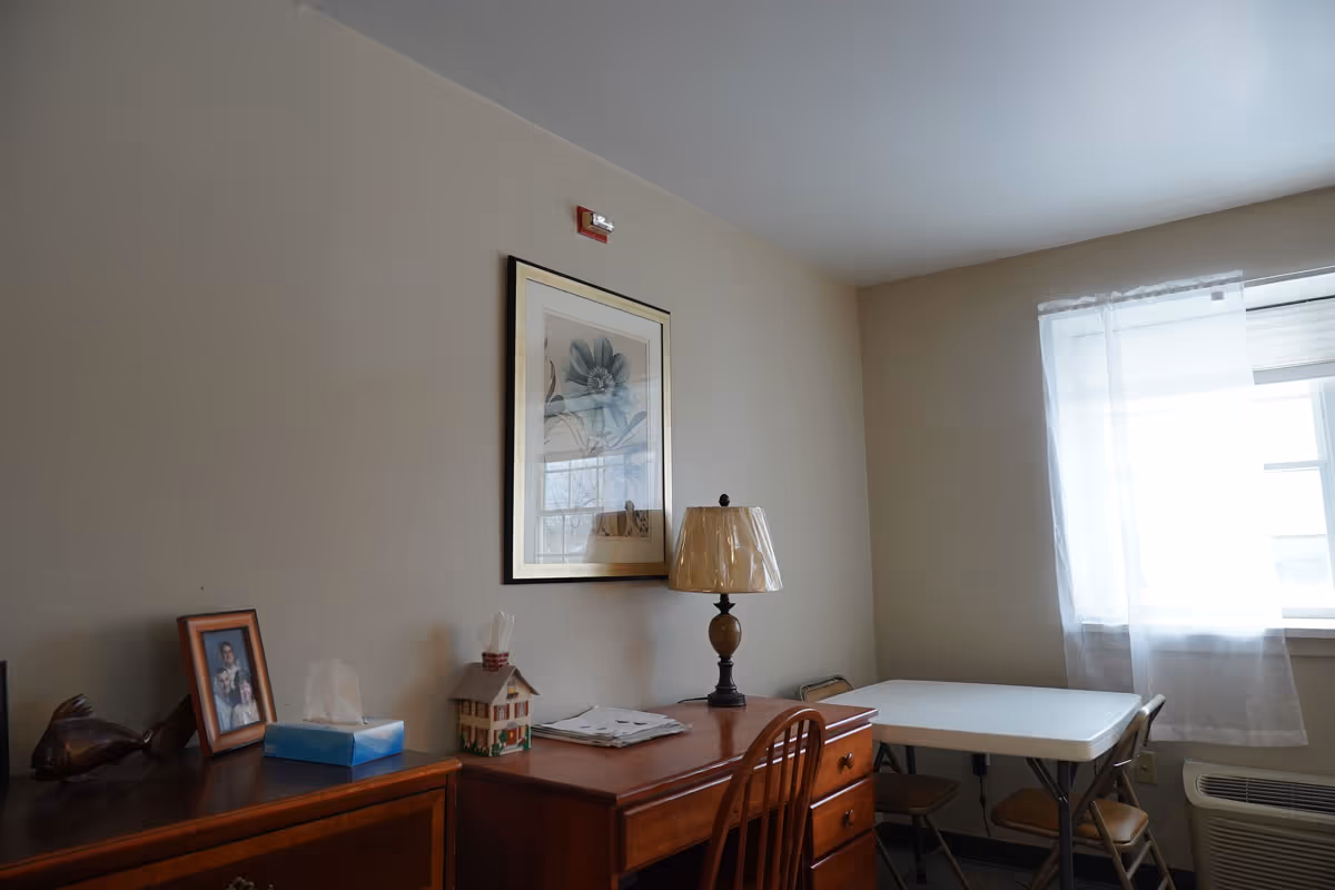 Small furnished interior with a wooden desk and dresser, lamp, framed artwork and photo, and a folding table with chairs by a curtained window.