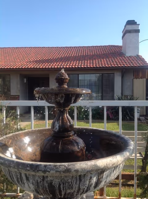 A weathered fountain sits on a railing with a single-story house in the background.