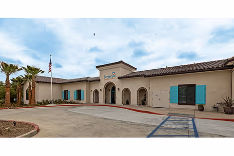 Exterior front view of Serra Sol Memory Care facility with beige stucco walls, turquoise window shutters, arched doorways, an American flag on a flagpole, palm trees, and a paved driveway with a handicapped parking space.