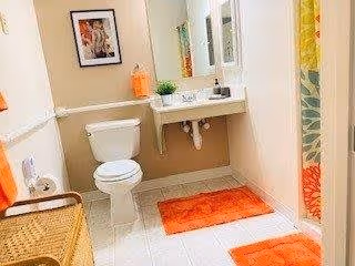 A clean bathroom featuring a white toilet, a wall-mounted sink with a mirror above it, and orange bath mats on the tiled floor. There is a wicker basket on the left side, a framed picture on the wall, and a shower curtain with a floral pattern on the right.
