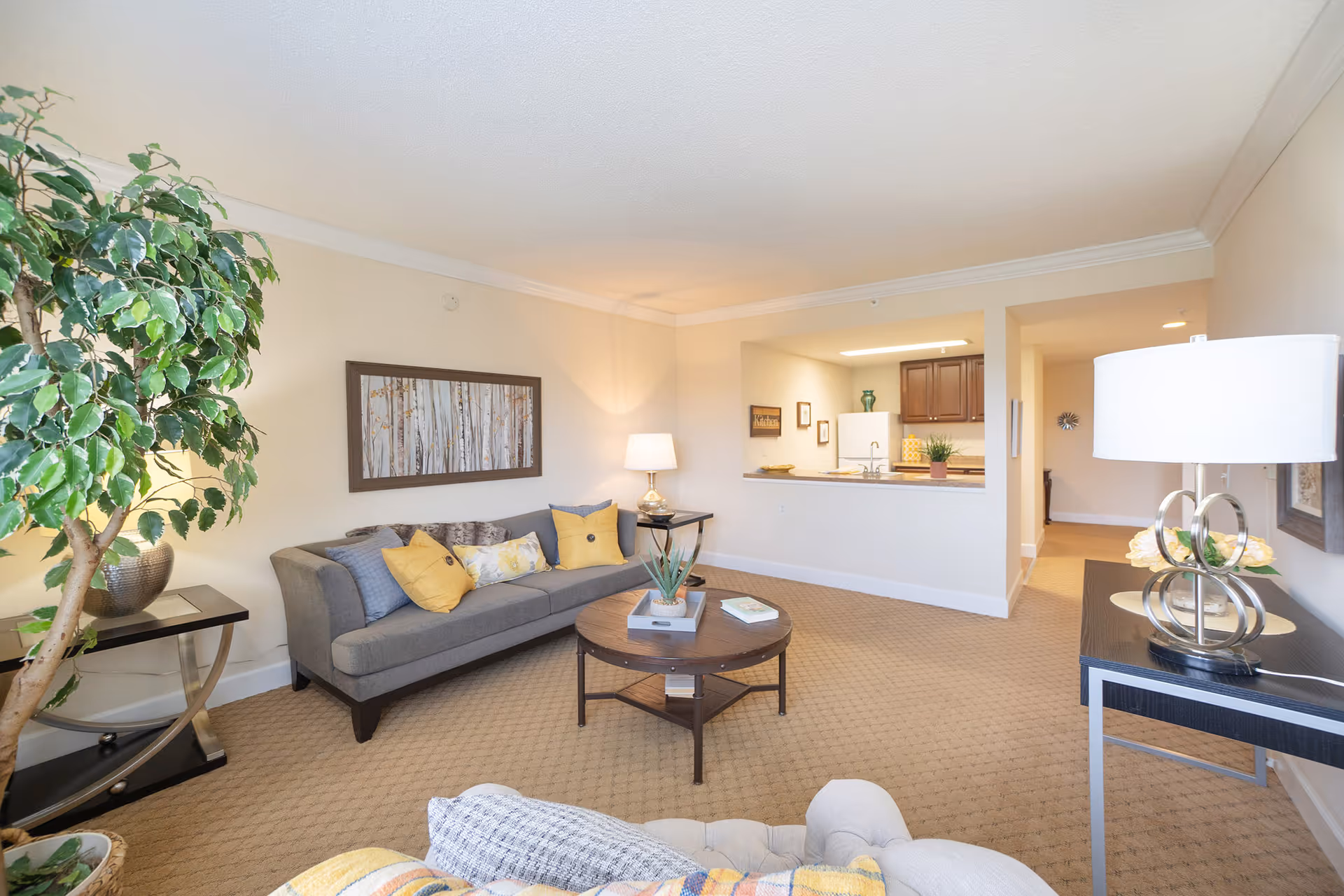 A bright and cozy living room with a gray sofa adorned with yellow and patterned cushions, a round wooden coffee table with books and a small plant, a large leafy indoor plant in a pot, a side table with a lamp, and a console table with a modern lamp and decorative items. The room has beige carpet and walls, and an open view into a kitchen area with dark cabinets and a white refrigerator.