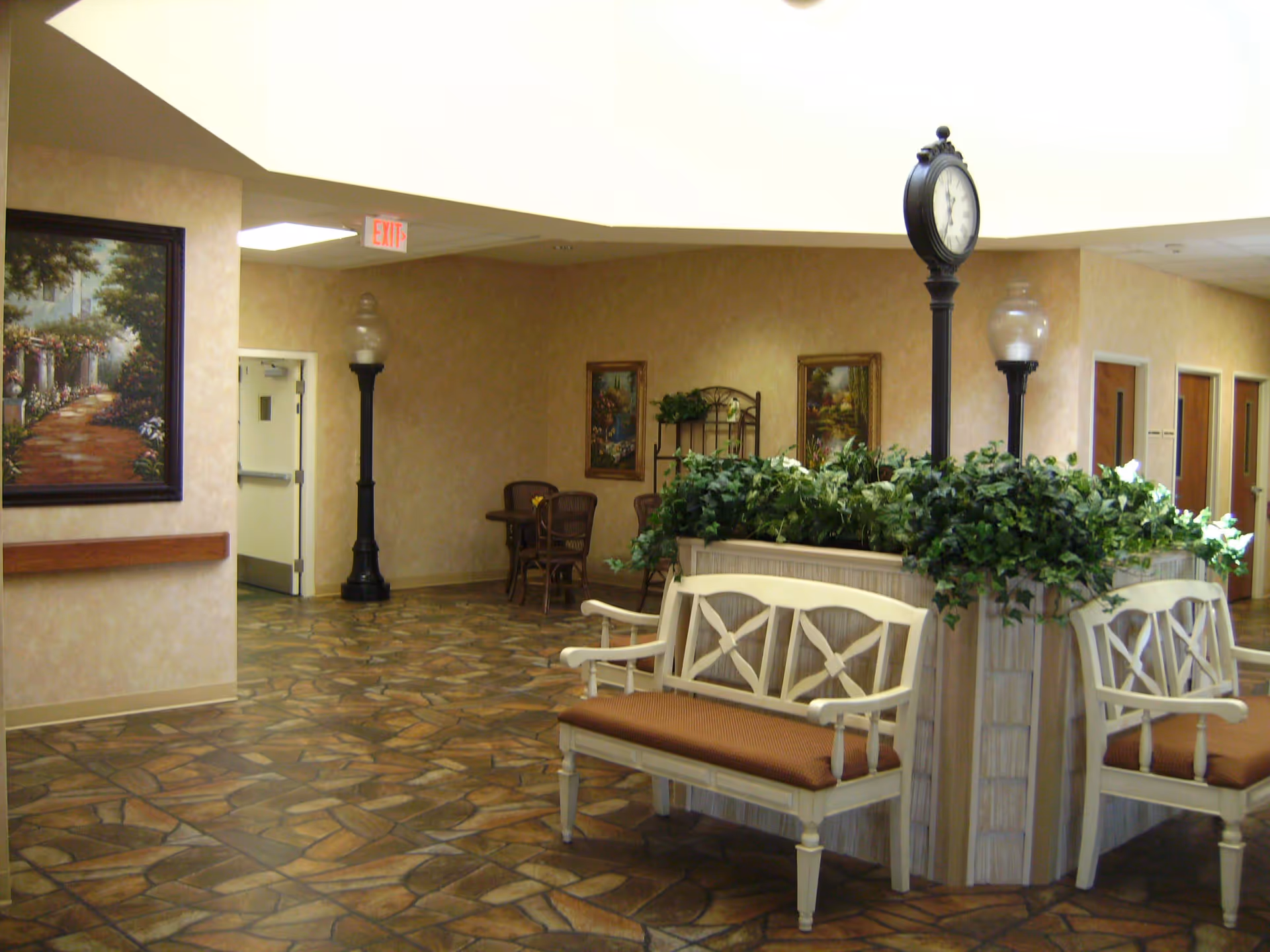 Interior common area of a senior living facility with beige walls and a patterned tile floor. There are two white wooden benches with brown cushions surrounding a planter filled with green plants. A tall black clock stands in the middle of the planter. In the background, there are two black lamp posts, framed paintings on the walls, a small table with chairs, and several doors. An exit sign is visible above a door on the left side.