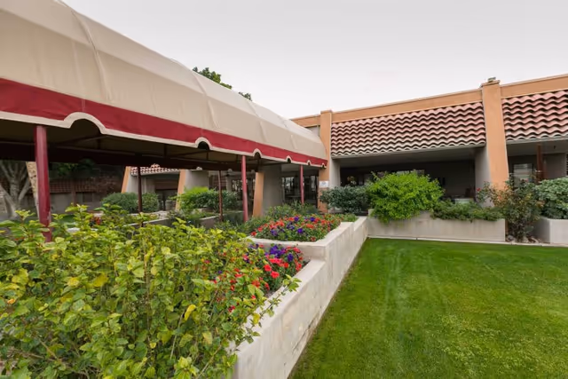 Outdoor garden area at Rio Vista Post Acute And Rehabilitation featuring a covered walkway with a beige and red awning, flower beds with colorful flowers, green bushes, and a well-maintained lawn in front of a building with a tiled roof.