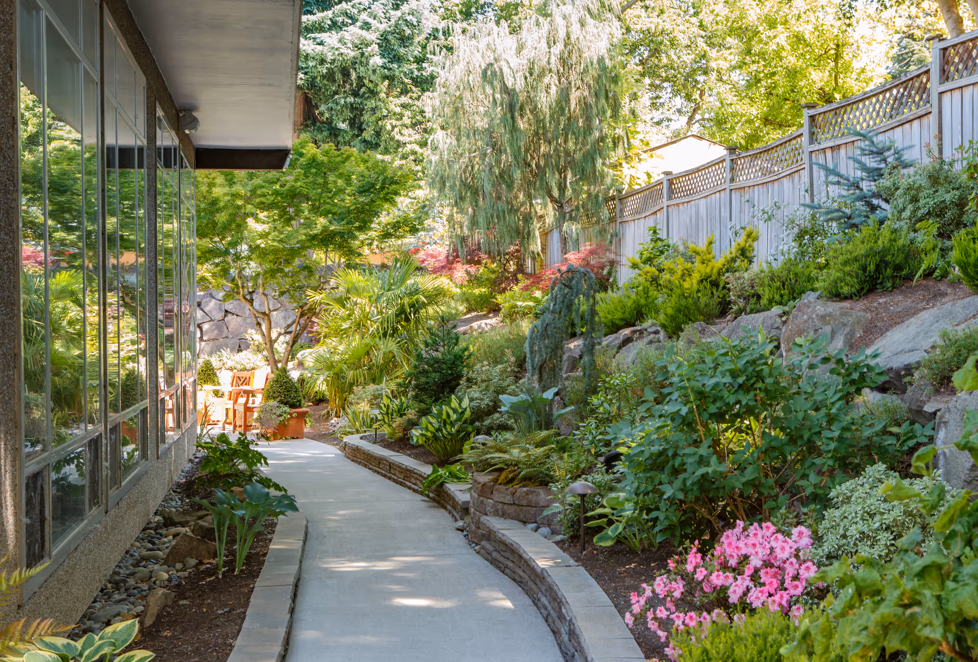 A paved walkway bordered by a variety of lush green plants and colorful flowers, leading alongside a building with large windows. A wooden fence runs along the right side, and there is outdoor seating visible in the background under trees.