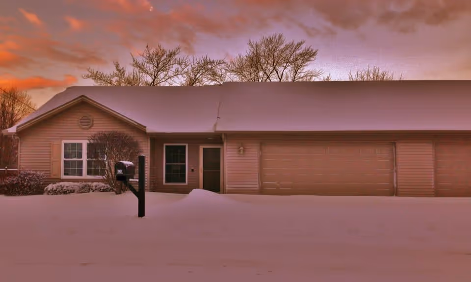 Exterior view of a single-story house covered in snow at sunset with a mailbox in the front yard and leafless trees in the background.