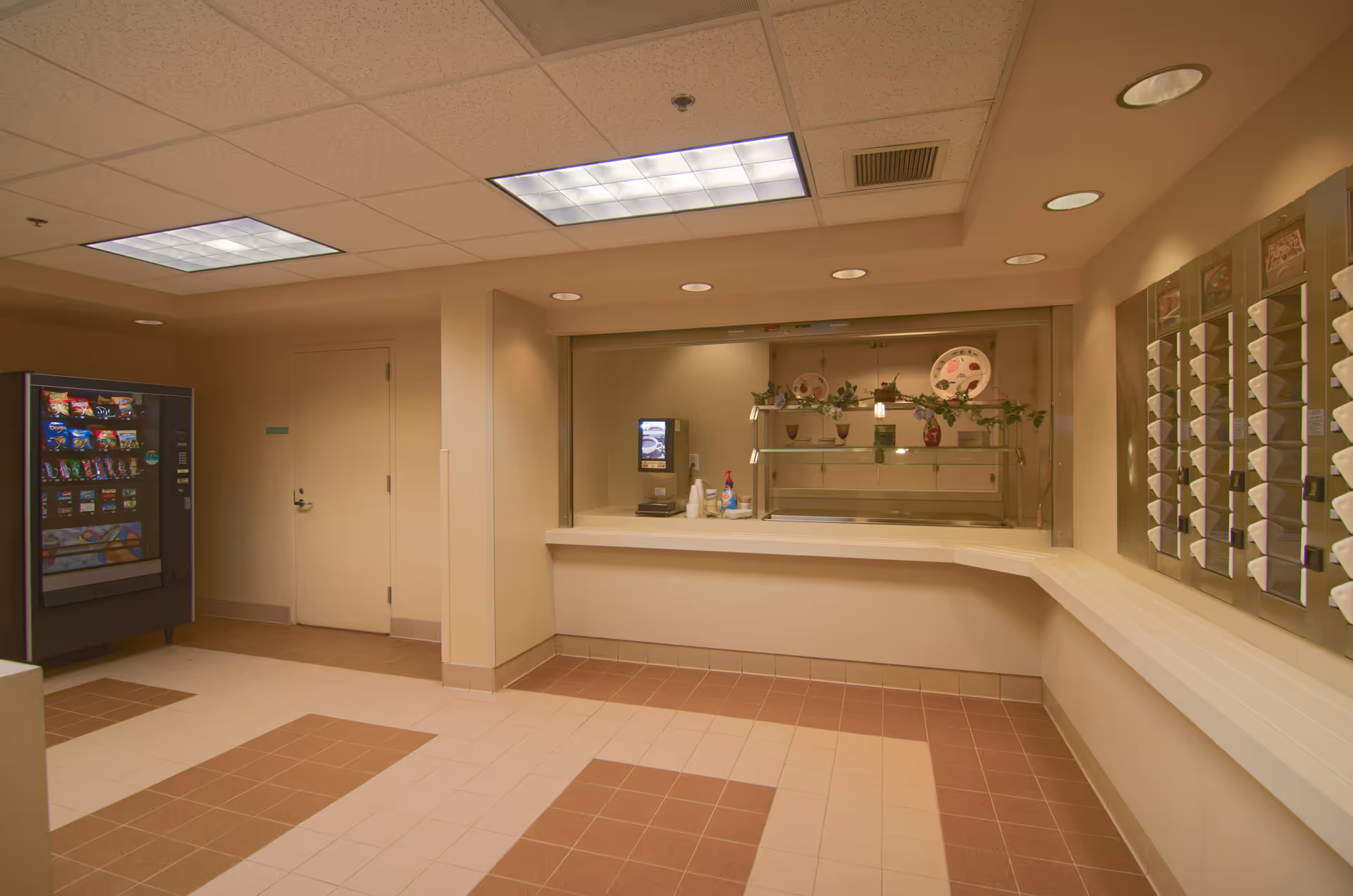 Interior view of a senior living facility snack and beverage area with a vending machine on the left, a counter with a beverage dispenser and cups in the center, and a wall-mounted mail or key holder on the right. The floor has a patterned tile design and the ceiling has recessed lighting.