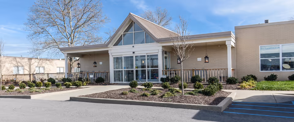 Front exterior view of Morgantown Healthcare Center building with a peaked roof entrance, glass doors, landscaped bushes, and a clear blue sky.